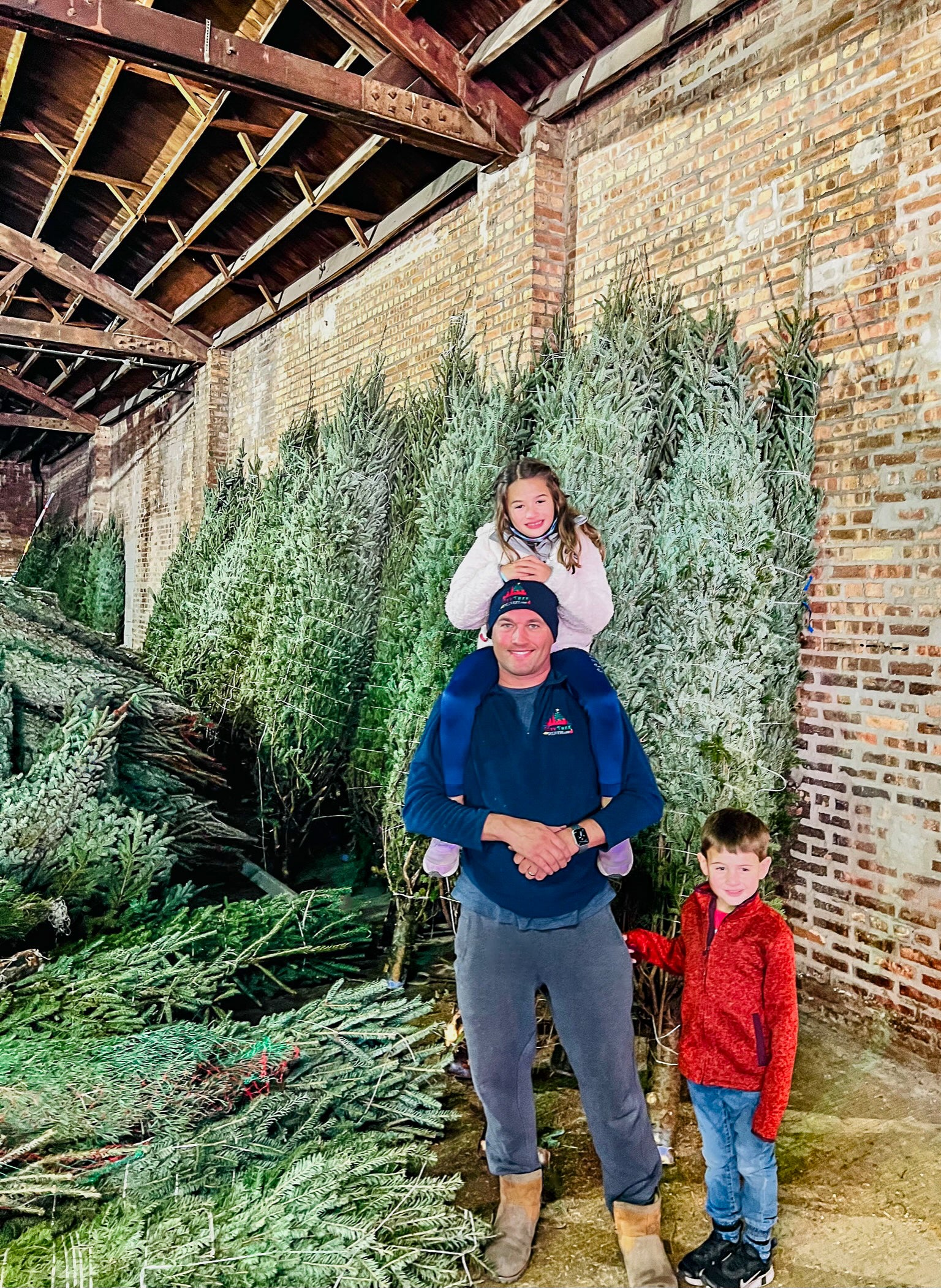 Family of three standing among Christmas trees in a rustic setting